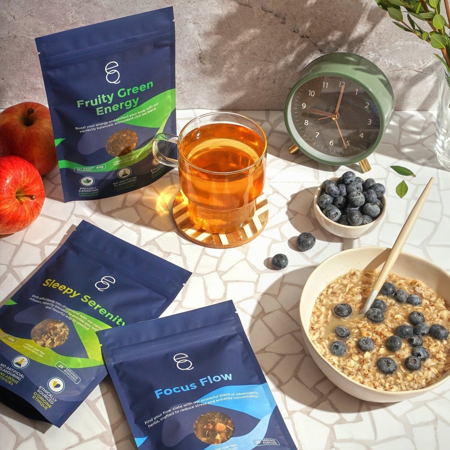 The image displays the All-Day Balance Gift Set, with three tea packages labeled 'Fruity Green Energy', 'Sleepy Serenity', and 'Focus Flow', alongside a cup of tea, a bowl of cereal with blueberries, an EQ Tea Spoon, EQ Tea Infuser, and a sand timer on a table with a clock, apples, and blueberries.