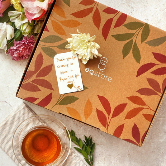 Tea box with floral design and a card, accompanied by a cup of tea and flowers on a light surface.