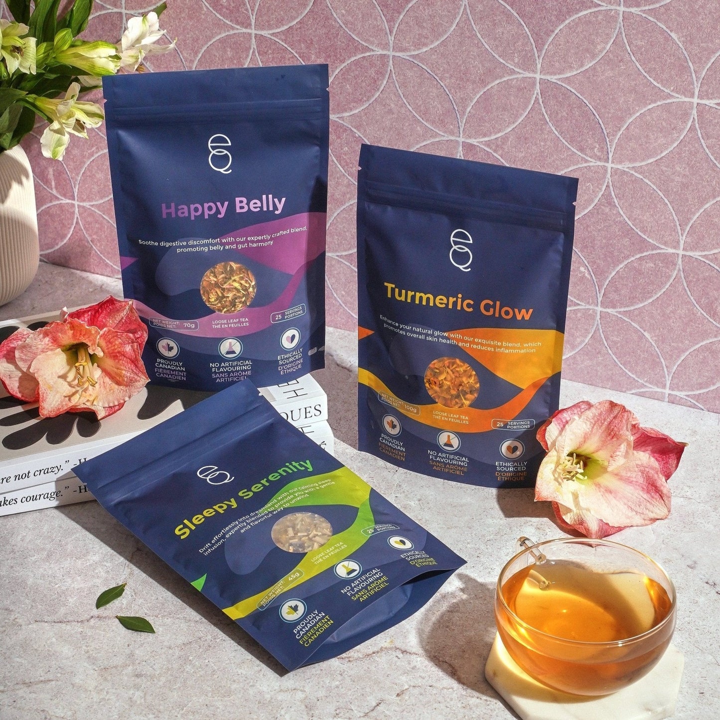 Product image of EQ Essentials Gift Set which includes tea boxes labeled 'Happy Belly', 'Turmeric Glow', and 'Sleepy Serenity', along with an EQ Tea Spoon, EQ Tea Infuser, and EQ Tea Timer, displayed on a table with a cup of tea and decorative flowers.