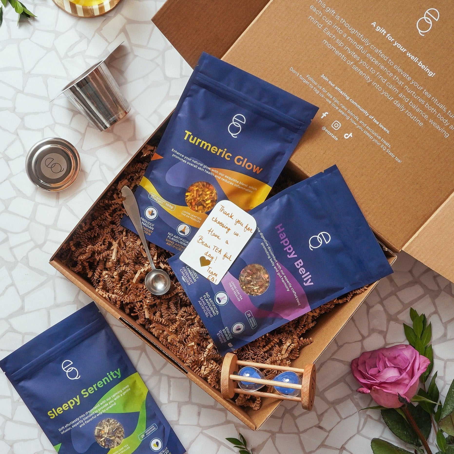 Box of 'Turmeric Glow', 'Happy Belly' and 'Sleepy Serenity' packages with a spoon and rose on a textured surface.